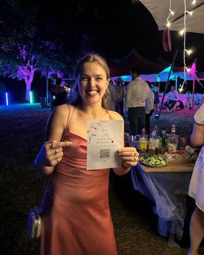 Eine lächelnde junge Frau (Trauzeugin) auf einer abendlichen Hochzeitsfeier hält eine Karte mit einem Hochzeit QR-Code in die Kamera und zeigt darauf, um andere Gäste zum Teilen ihrer Fotos und Erinnerungen zu animieren.