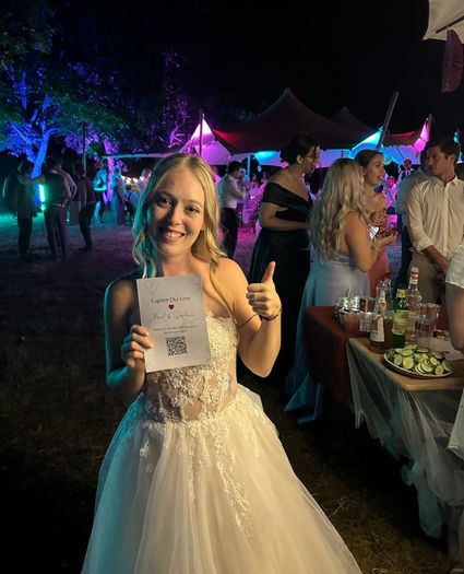 Die glückliche Braut hält bei ihrer abendlichen Hochzeitsfeier eine Karte mit einem Hochzeit QR-Code in der Hand und gibt einen Daumen nach oben, um ihre Gäste zum digitalen Teilen von Fotos zu motivieren.
