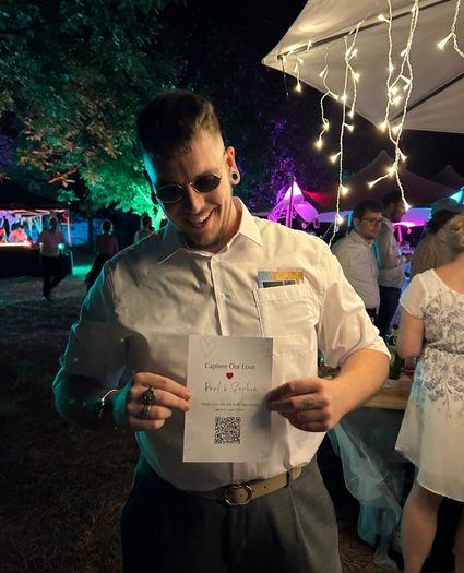 Ein lächelnder Gast oder DJ auf einer Hochzeitsfeier im Freien hält eine „Capture Our Love“-Karte mit einem Hochzeit QR-Code hoch, um die Gäste auch während der Party zum Hochladen von Schnappschüssen zu animieren.