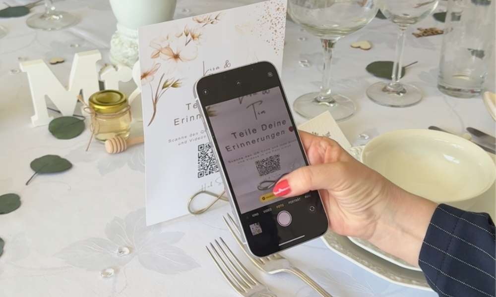 Ein Gast scannt mit einem Smartphone einen Hochzeit QR-Code auf einer eleganten Tischkarte, um Fotos der Hochzeitsfeier direkt in das digitale Album hochzuladen.